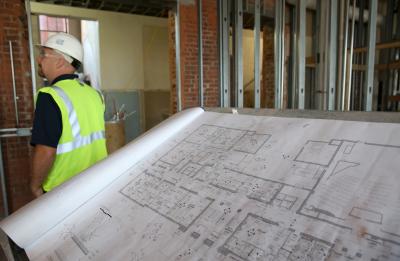 Man wearing a safety vest and hardhat stands next to renovation plans.