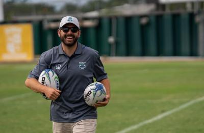 A person wearing a dark gray staff polo shirt and khaki shorts walking on a rugby field while carrying two rugby balls, one in each arm. The background shows field equipment and fencing.