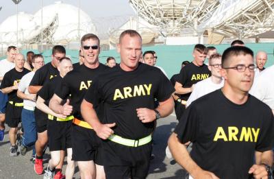 A group of people wearing black ARMY physical training shirts runs together outdoors near a secure facility with satellite dishes and barbed-wire fencing in the background.