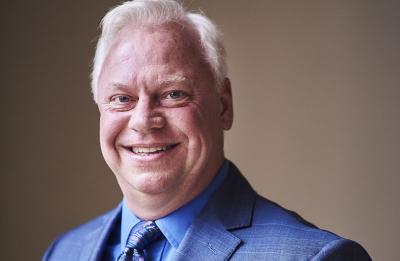 A person wearing a blue suit jacket, blue shirt, and patterned tie is photographed in a professional portrait setting. The background is softly lit with neutral tones, and the subject is framed from the chest up.