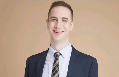 A person wearing a dark blazer, patterned tie, light-colored dress shirt, and brown belt stands against a plain beige background. The individual is posed with hands in pockets, and the outfit is neatly arranged and professional in appearance.