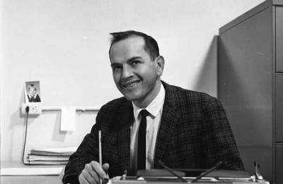 Black-and-white photo of a person seated at a desk, holding a pencil beside a typewriter, with papers and office furniture visible in the background.