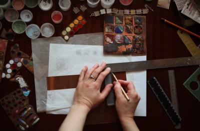     Query successful  Gemini said  An overhead view shows an artist’s hands using a metal ruler and a stylus to mark a copper sheet on a cluttered workbench. The workspace is surrounded by various tools of the trade, including colorful enamel powders in small containers, metal palettes with fused glass samples, and several rulers.