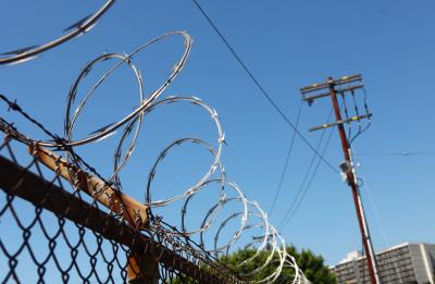 azor wire coiled on top of a chain-link fence against a clear blue sky, with a utility pole and urban buildings visible in the background.
