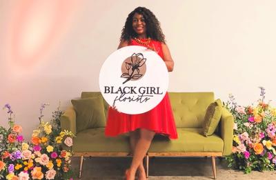  Person in a red dress stands in front of a green couch holding a round sign that reads “Black Girl Florists,” with floral arrangements on both sides.
