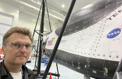 A person stands inside a large industrial facility next to a spacecraft or rocket component covered in black and white thermal panels, with a NASA logo and U.S. flag visible on the surface. Support structures and equipment surround the vehicle.