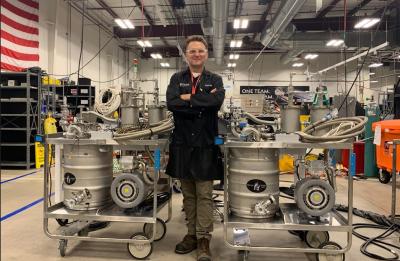 A person stands with arms crossed between two stainless steel vacuum cart ground support equipment units on wheeled frames inside an industrial aerospace facility. Each cart has hoses, valves and gauges, with high ceilings, ductwork and an American flag visible in the background.