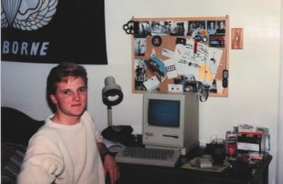 A person sits at a desk in a dorm-style room with an early desktop computer and keyboard, surrounded by a desk lamp, posters, and personal items pinned to the wall.