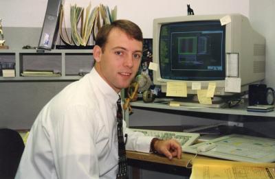 A person seated at a desk in an office with an early computer monitor displaying a technical design, surrounded by paper files and sticky notes.