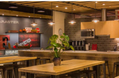 A modern office kitchen and lounge area with wood tables, bar stools, pendant lighting, and wall décor featuring words like “Community” and “Character.”