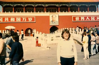 A person stands in a public square near a large red building with Chinese signage, with a crowd of people in the background.