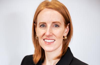 Person with shoulder-length red hair wears a black blazer and gold earrings, posed against a light gray background.