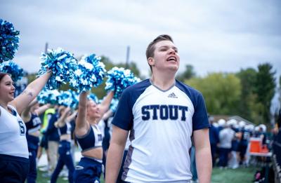UW-Stout student in a “STOUT” shirt cheers on the sidelines while a group of cheerleaders raise blue pom-poms at a football game.
