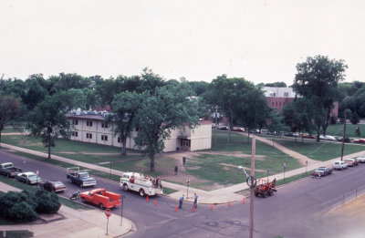 Aerial view of a campus corner with a low-rise academic building surrounded by mature trees and lawns. Several cars are parked along the street while utility workers use a truck and cones to work near an intersection, with sidewalks and paths crossing the green space.