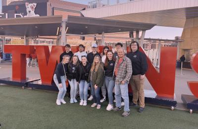 Group of students and one adult pose in front of large red “TWINS” letters outside a stadium.