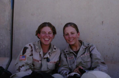 Two people in U.S. Army uniforms sit side by side outdoors against a concrete wall with blue stenciled text, smiling at the camera.