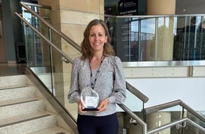 Person standing on an indoor staircase holding a small rectangular award plaque, with glass railings, beige columns and a campus-style atrium visible in the background.