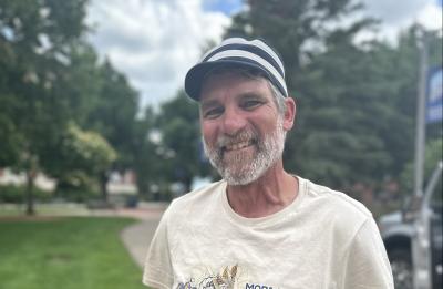 Portrait of a mural artist standing outdoors near a sidewalk and grassy area wearing a paint-splattered T-shirt.