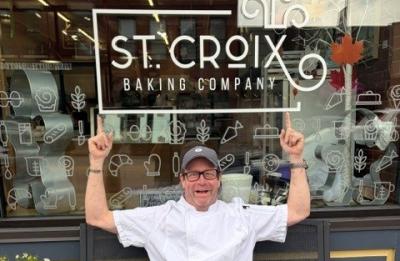 A person wearing a white chef coat, tan pants and a gray cap sits on a black bench outside St. Croix Baking Company with both arms raised and pointing toward the bakery sign in the window behind them.