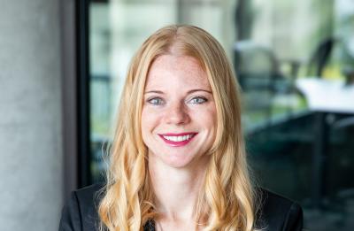 Professional headshot of a person with long light-colored hair wearing a dark blazer, seated indoors.