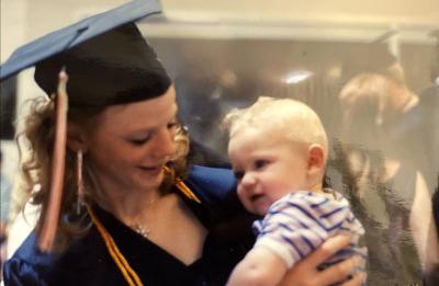 Graduate in a cap and gown holding a baby indoors during a commencement celebration.