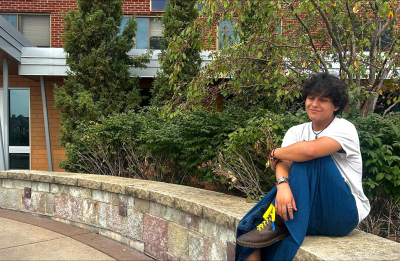 Person wearing a white T-shirt, blue pants and brown boots with bright yellow laces sits on a curved stone wall outside a brick building. Shrubs, evergreen trees and a leafy tree with small red berries fill the background, while a sidewalk curves through the foreground.