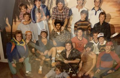 Large group of students posing for a residence hall group photo indoors in front of a tropical beach-themed backdrop. The group is arranged in several rows, with some students standing, sitting or kneeling, and many wearing casual clothing, costumes and hats. Several hats display the word “Stout.”