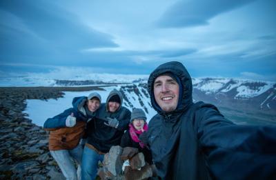 Four students in winter clothing pose for a selfie on a rocky mountain ridge overlooking snow-covered peaks and a remote landscape in Iceland.