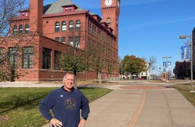 Poul David “Dave” Rasmussen stands on a campus walkway in front of UW-Stout Polytechnic's Bowman Hall and clock tower on a sunny fall day.