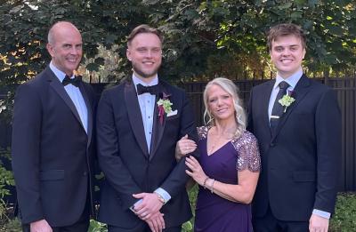 Four family members dressed in formal attire pose outdoors on a lawn in front of trees and landscaping during a wedding celebration.