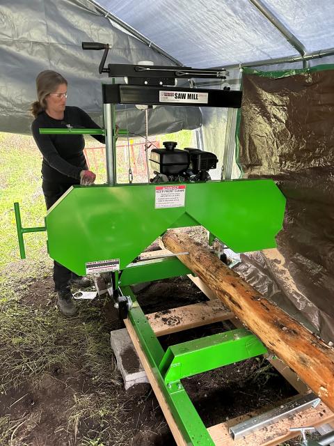 Individual standing beside a bright green sawmill under a tarp-covered workspace, cutting a log placed on the machine’s track.
