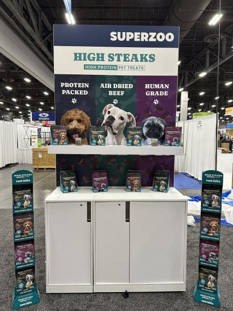 Trade show display for High Steaks high‑protein pet treats at the SuperZoo expo. The booth features a large sign with the brand name and three sections labeled ‘Protein Packed,’ ‘Air Dried Beef,’ and ‘Human Grade,’ each with a dog photo above rows of packaged pet treats. The setup includes a white counter, product samples arranged on shelves, and two branded floor stands on either side. The background shows an indoor convention hall with booths and equipment.