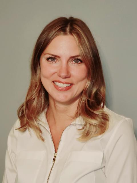 Person with shoulder-length wavy hair wearing a light-colored zip-up blouse, posed against a plain gray background.
