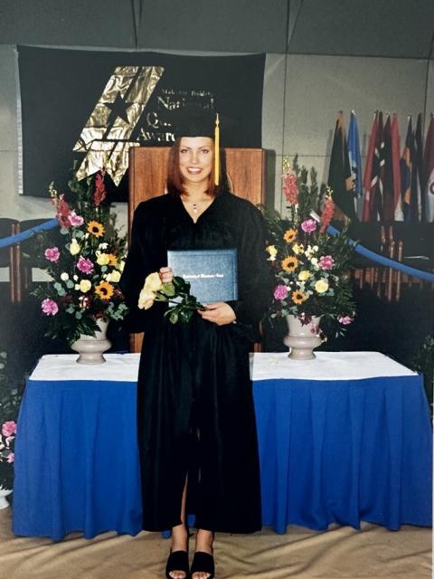 Person wearing a black graduation gown holding a diploma and flowers on a decorated commencement stage with floral arrangements and international flags in the background.