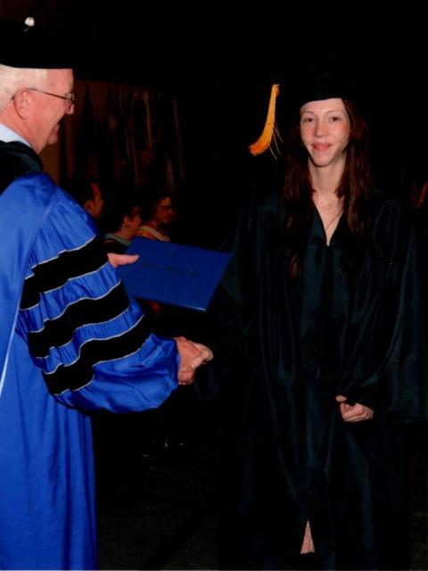 Graduate in a black cap and gown shaking hands with a person in a blue academic robe while receiving a diploma during a commencement ceremony.