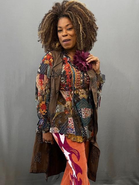 A person is wearing a vibrant, eclectic outfit with a strong bohemian and retro aesthetic. The top is a patchwork-style garment featuring a mix of colorful patterns, including florals and geometric designs, layered under a long brown coat adorned with intricate embellishments. A large purple flower accessory is pinned near the neckline, adding a bold accent. The pants are striking orange bell-bottoms with dramatic flame-like appliqué designs in shades of pink, white, and red running down the legs. 