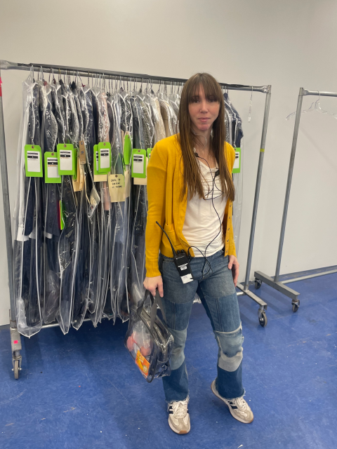 A person stands in a wardrobe workspace holding a transparent set bag. They are wearing a yellow cardigan, white shirt, and patchwork denim pants with sneakers. Behind them is a rolling rack filled with neatly organized costumes in garment bags, each tagged with bright green labels. The setting appears to be a production or costume department area with blue flooring and additional empty clothing racks nearby.