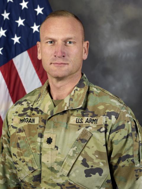 A person wearing a U.S. Army camouflage uniform poses for a formal portrait in front of an American flag and neutral studio backdrop.