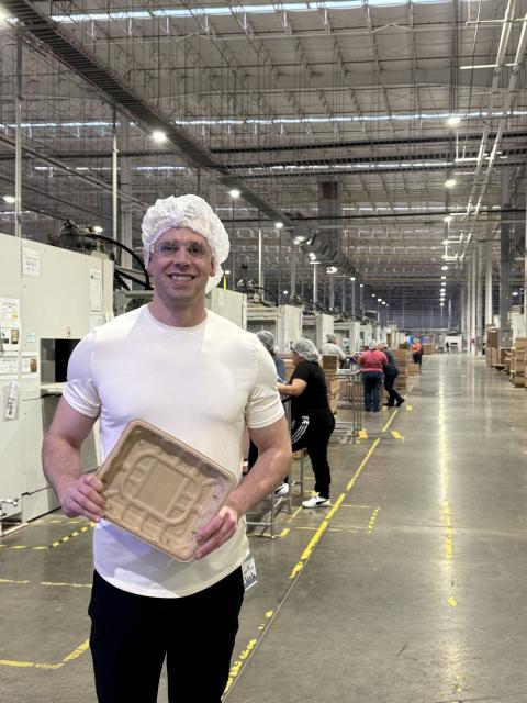 A person wearing a white T-shirt and a hairnet stands in a brightly lit industrial facility, holding a molded fiber food tray. The factory floor extends into the distance with machinery, workstations, and other workers assembling or inspecting products.