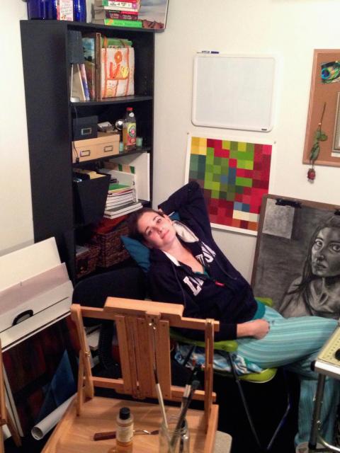 art studio with paintings, sketches and supplies throughout the space. An easel with paintbrushes sits in the foreground, while several canvases lean against the wall on the left. A tall black bookshelf holds art materials, books and storage boxes. A person reclines in a chair wearing casual clothes, surrounded by artwork and a laptop workspace on a table to the right. The walls display corkboards with inspiration images, a color grid print and a blank whiteboard.