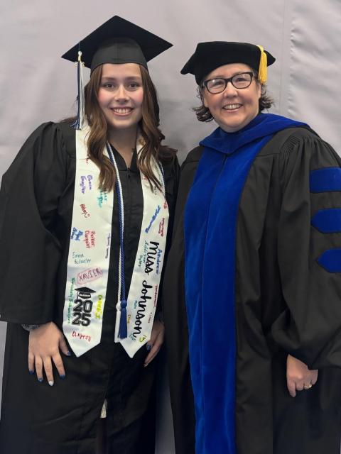 Two individuals wearing academic regalia stand side by side. One is dressed in a graduation cap and gown with a decorated stole featuring handwritten messages. The other is wearing a doctoral robe with a blue velvet panel and a tam with a gold tassel. They are standing in front of a plain background.