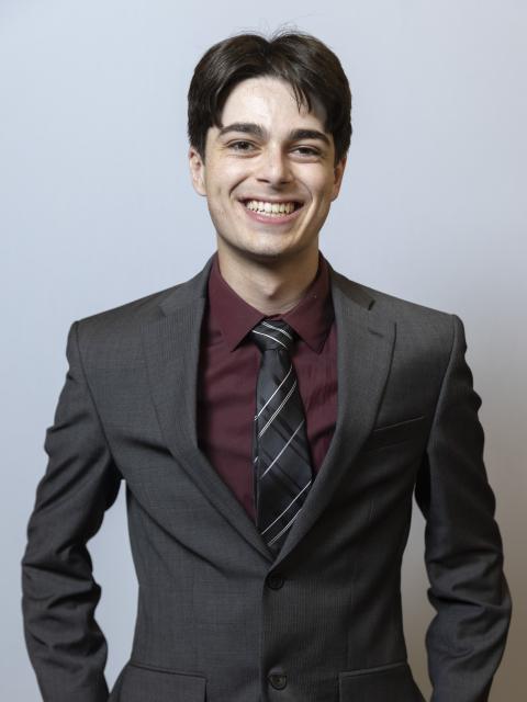 A person stands against a light gray background wearing a dark gray suit, maroon dress shirt and striped tie, facing the camera with hands in pockets.