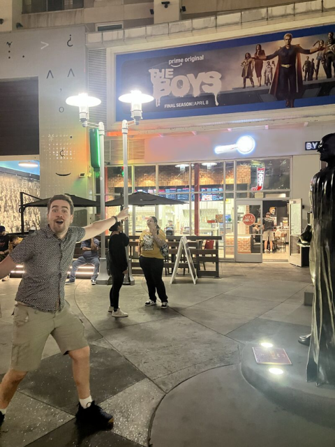 Michael Hilliger points toward a billboard for The Boys while standing in an outdoor entertainment district at night.