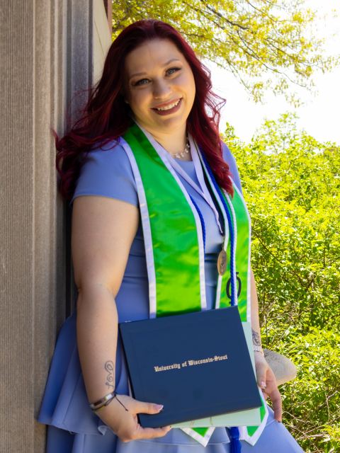 UW-Stout graduate posing outdoors while holding a diploma and wearing graduation cords, a medallion and a green stole.