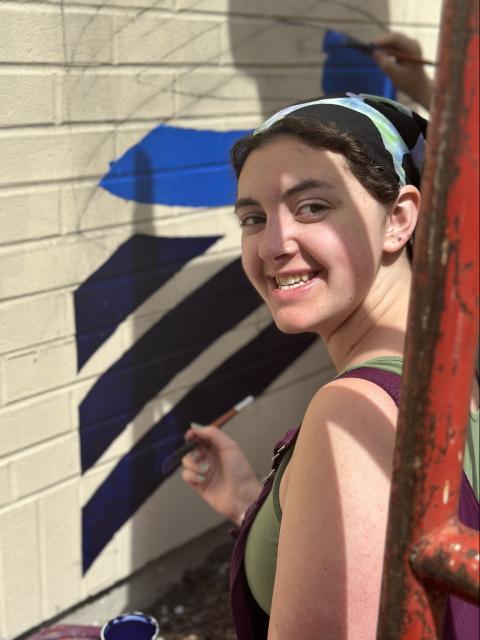 Person painting blue geometric shapes on a brick wall mural while looking toward the camera.