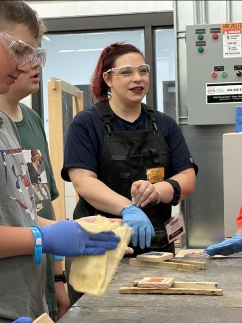 Instructor wearing safety glasses and gloves demonstrates a woodworking finishing technique to students gathered around a worktable.