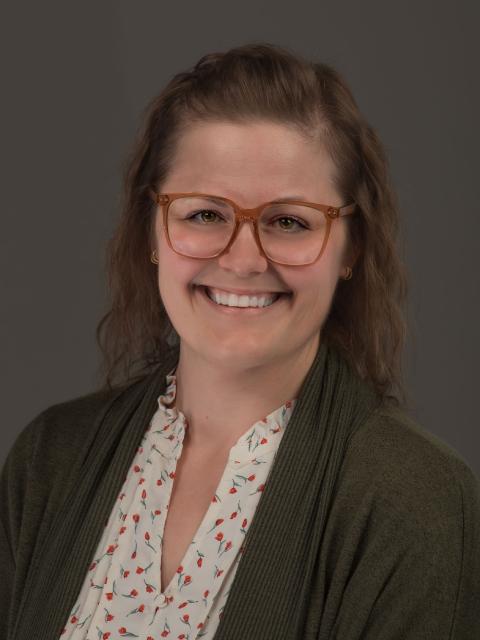 Smiling woman with glasses, wavy brown hair, wearing a patterned blouse and green cardigan against a plain background