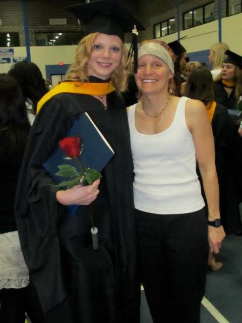 Two people pose together at a graduation ceremony. One person wears a black graduation gown and cap, holding a diploma cover and a red rose, while the other wears a white sleeveless top and dark pants. Other graduates and attendees are visible in the background of the gymnasium.