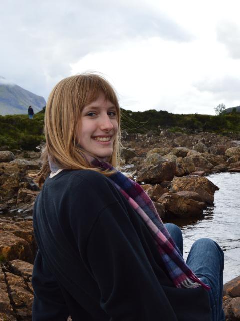 Julia sits on rocks beside a flowing river with mist-covered mountains and cloudy skies in the background.