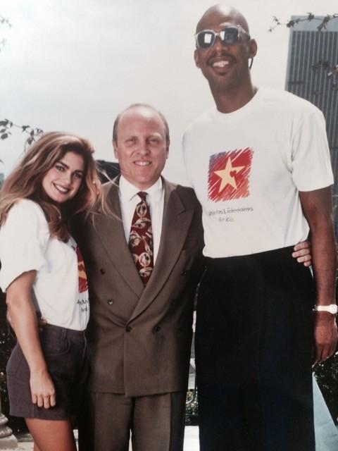 Historic photo of three people standing together outdoors. One person in a suit stands between two people wearing matching event T-shirts.